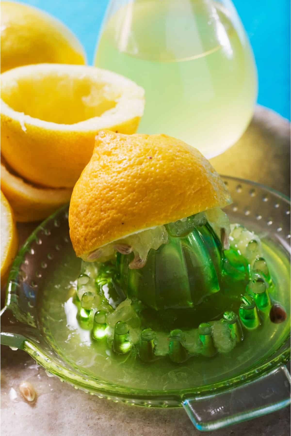 A green handheld lemon juicer with a half of a lemon being juiced on top of it. In the background are already juice lemon halves an a small jug of lemon juice.
