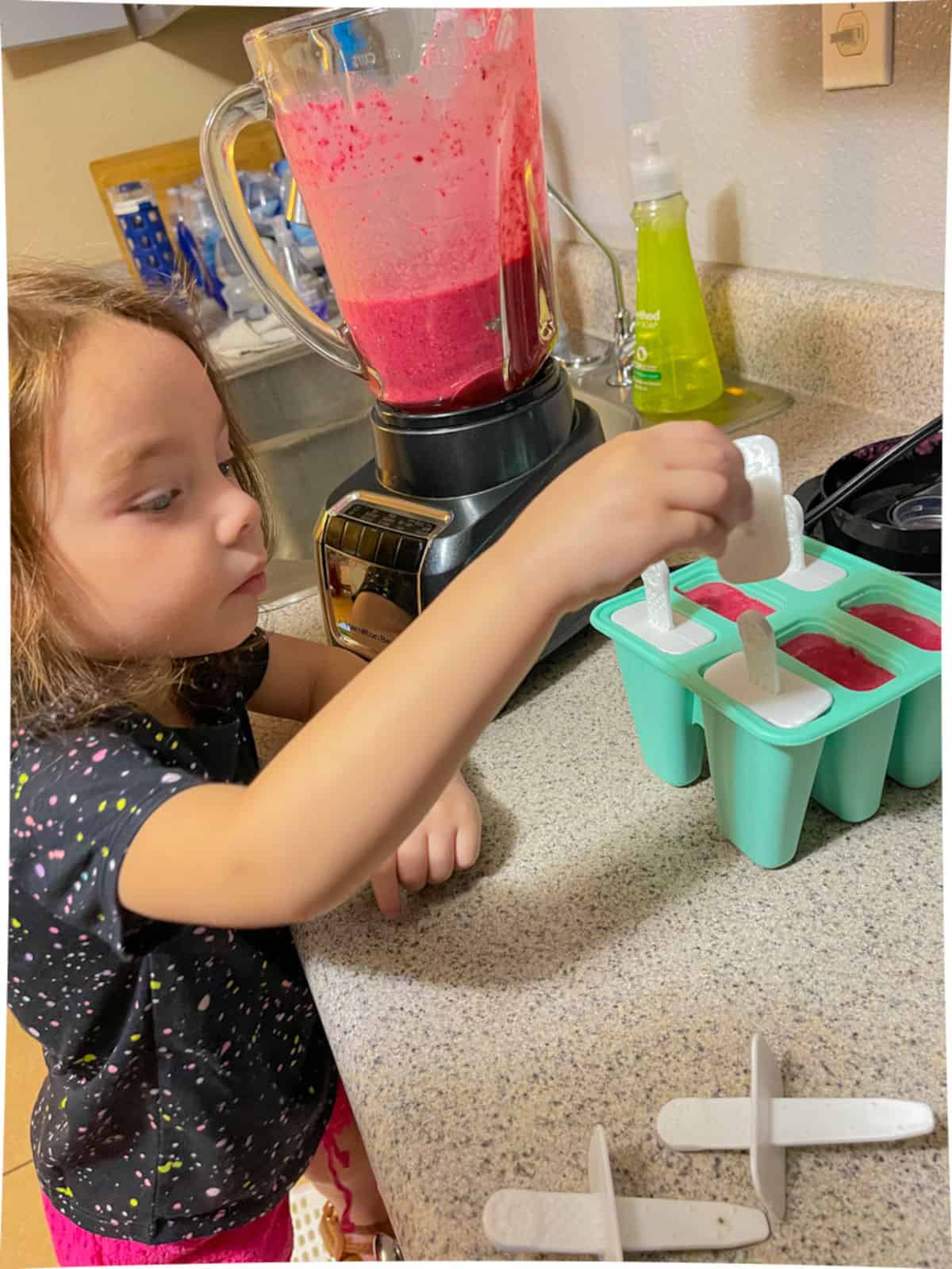 A small child wearing a black shirt with multicolored paint spray spots and pink shorts. She is standing on a step stool at the kitchen counter with a teal popsicle mold next to a blender filled with strawberry colored juice in it. The juice is being filled into the popsicle mold and there arer white sticked the child is placing into the mold.
