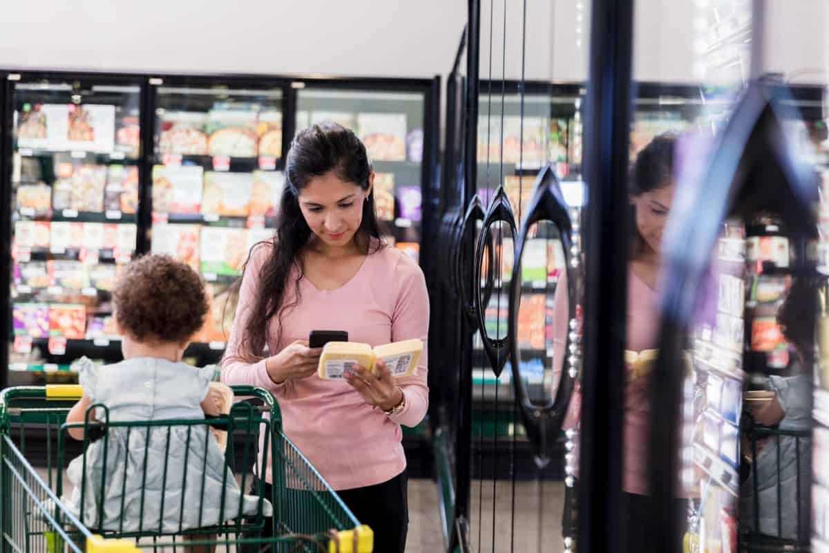 A woman with a baby in a grocery shopping cart comparing two products with her phone out.