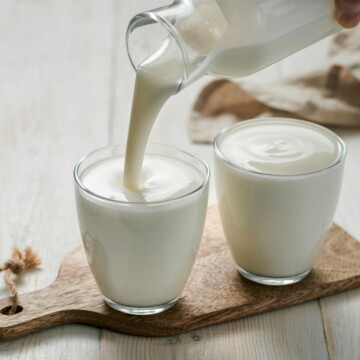 A cutting board with two glasses on it filled with kefir being poured from a larger carafe