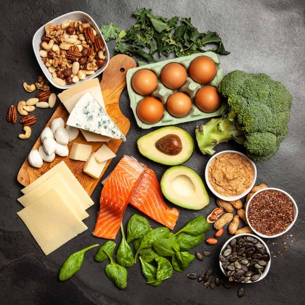 Clean keto ingredients on a table: spinach, seeds, nuts, fish, cheese, eggs, avocado, nut butter, broccoli, and arugula.
