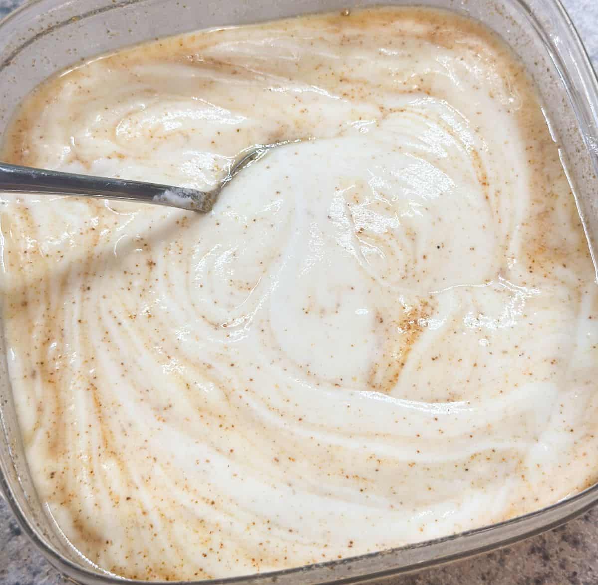A close up of a dish wish just mixed sweet and spicy yogurt sauce lightly yellow with specs of cayenne pepper in it and a spoon.