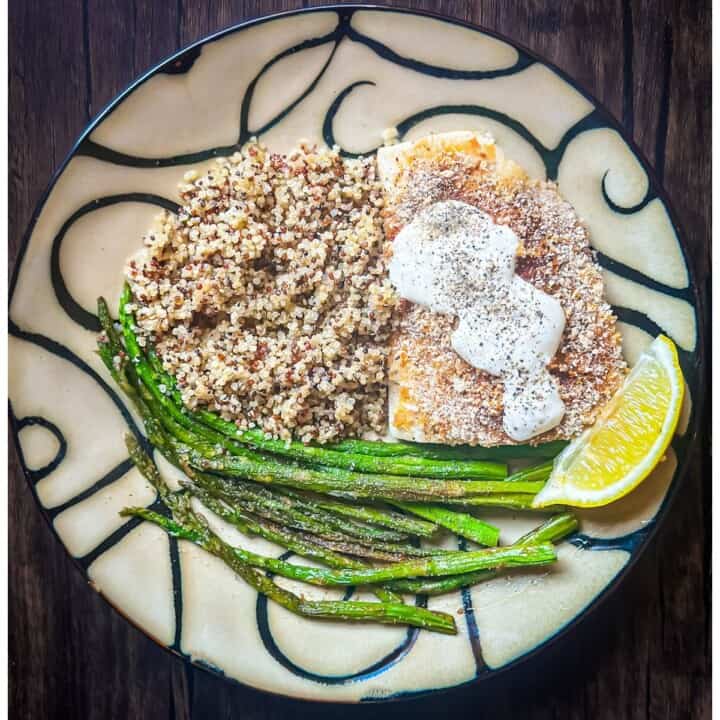 A tan and black swirled decorative round plate with a Mahi Mahi fillet covered in lightly browned bread crumb and spice mixture and topped with spicy yogurt sauce that was seasoned with black pepper. Next to the fish is a lemon wedge, a pile of multi-colored quinoa, and a side of sautéed asparagus.