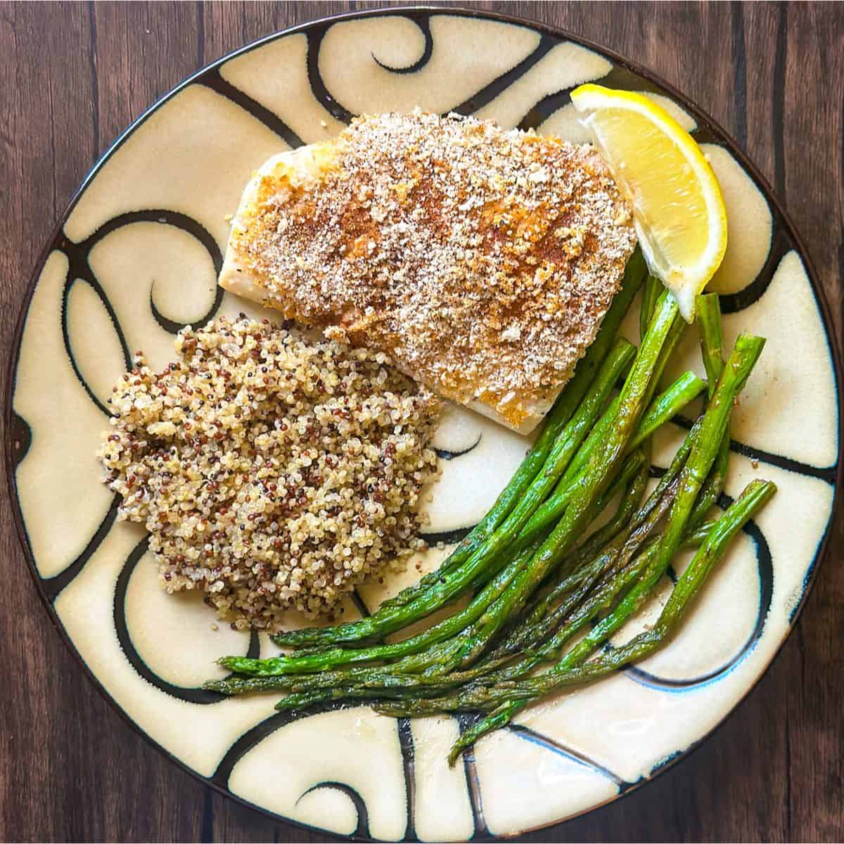 A tan and black swirled decorative round plate with a Mahi Mahi fillet covered in lightly browned bread crumb and spice mixture. Next to the fish is a lemon wedge, a pile of multi-colored quinoa, and a side of sautéed asparagus.