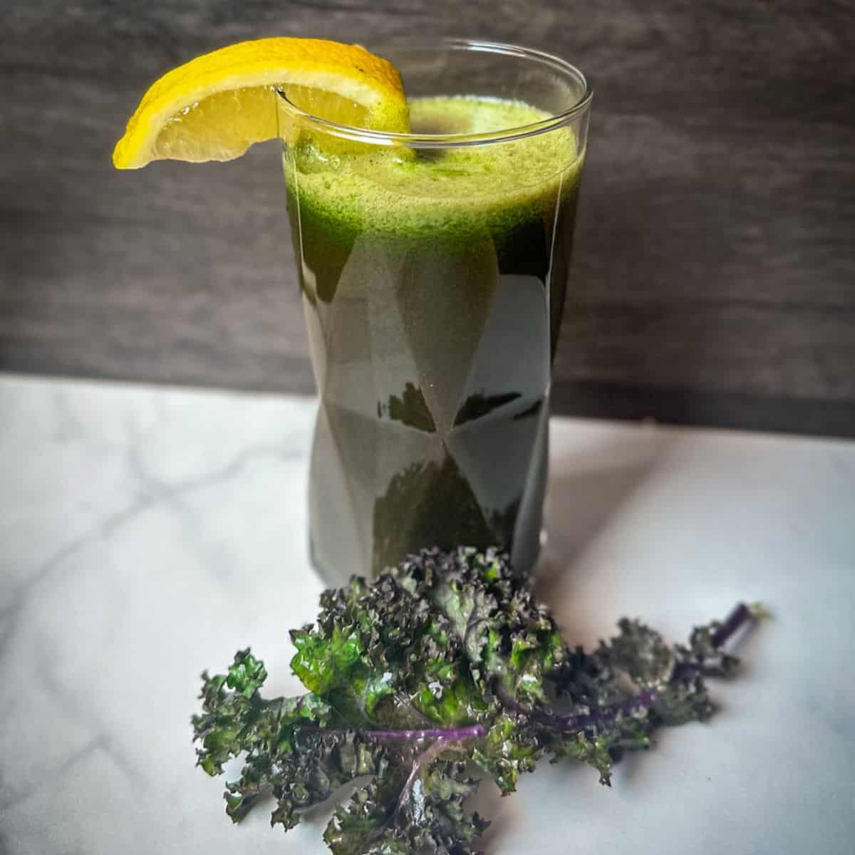 First Watch Kale Tonic Green Juice in a glass with light green foam at the top. The glass is garnished with a lemon wedge and a kale leaf at the base.