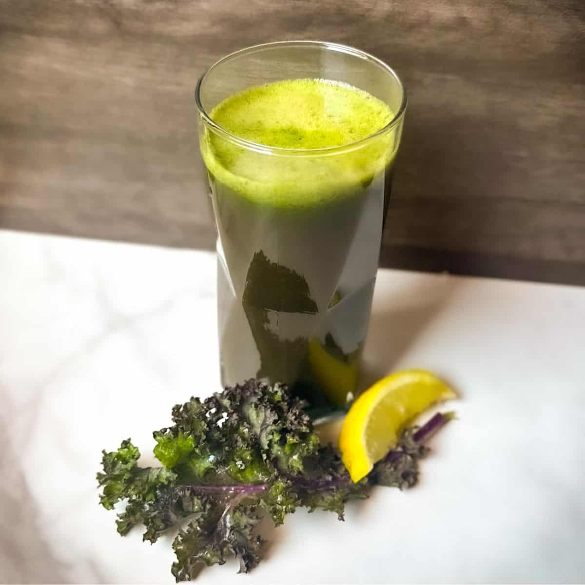 A clear tall glass of green juice (kale tonic) with a kale leaf and lemon wedge at the base for garnish.