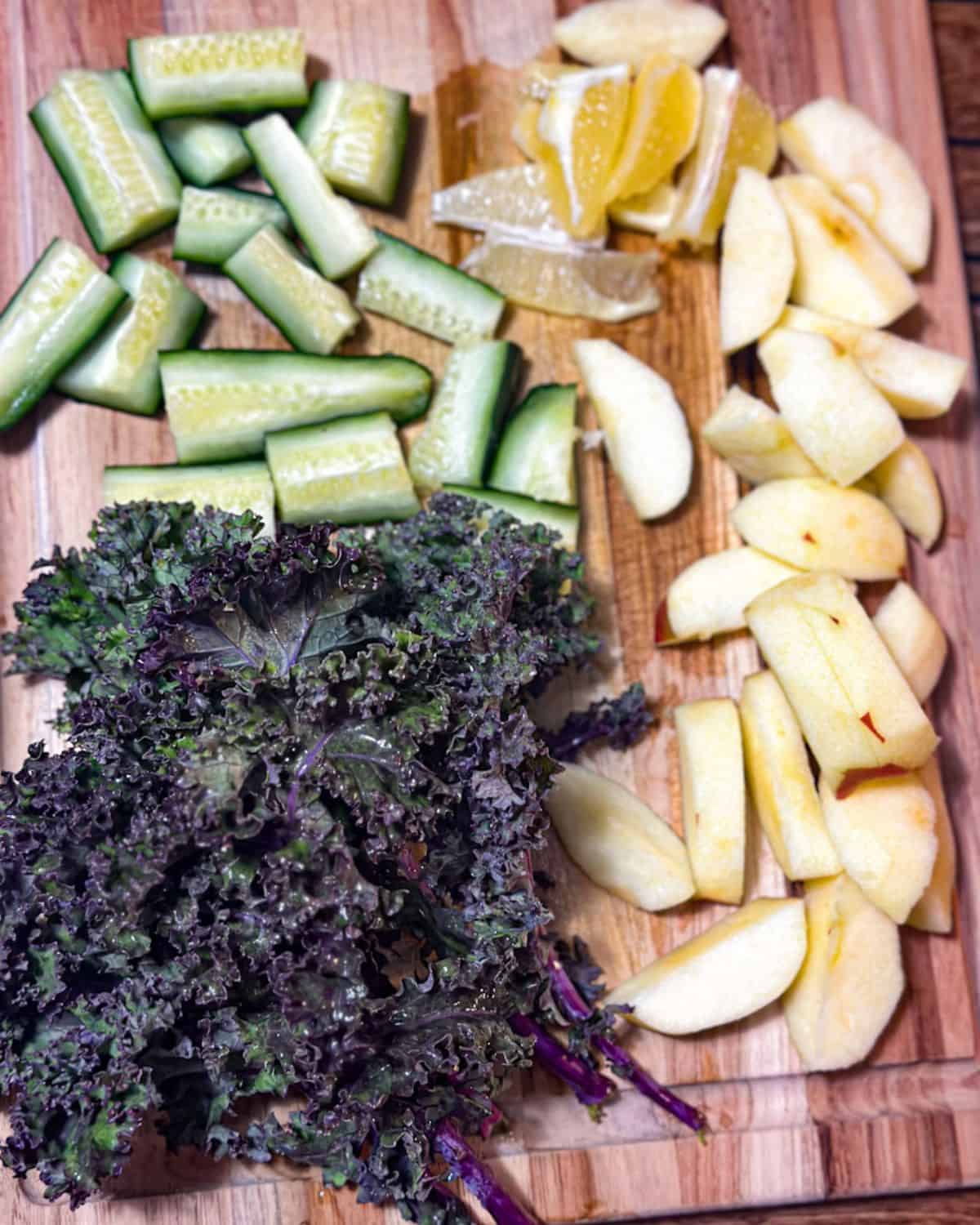 Purple kale leaves, chopped apples, chopped cucumbers, and chopped lemon without the rind sit on a wooden cutting board.