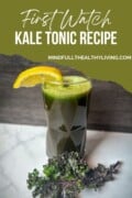 A torn-looking photo of green juice kale tonic with a lemon wedge garnish and a kale leaf at the base. Text overlay says first watch kale tonic recipe mindfullyhealthyliving.com