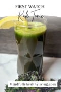 A green juice kale tonic in a clear glass with a lemon wedge and kale leaf that fades to white at the top. Text overlay says first watch kale tonic mindfullyhealthyliving.com