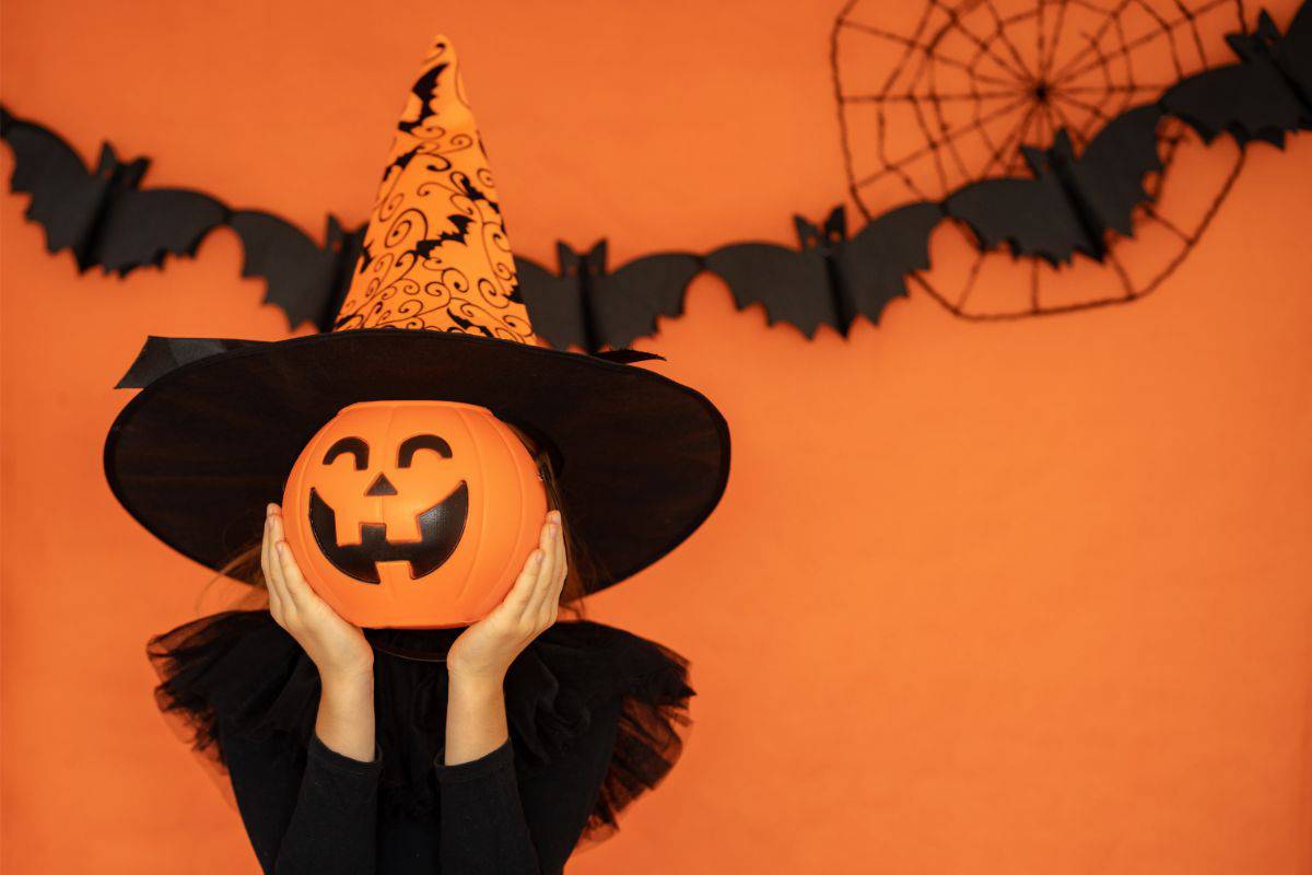 A photo of a child in a black dress and orange and black witch's hat holding a plastic pumpkin bucket in front of their face. The background is orange with black bats decoratively cut with a black spiderweb on the wall behind them.