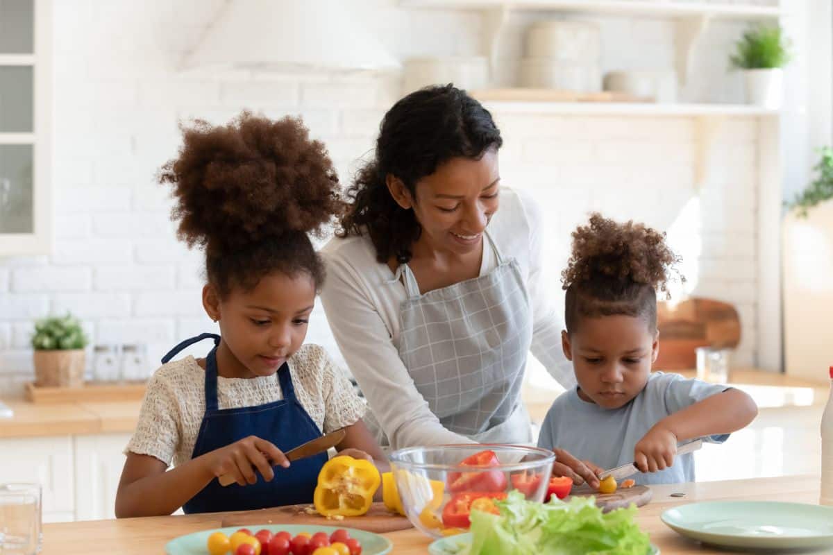 A woman with two young children in the kitchen learning to prepare colorful fruits and vegetables.