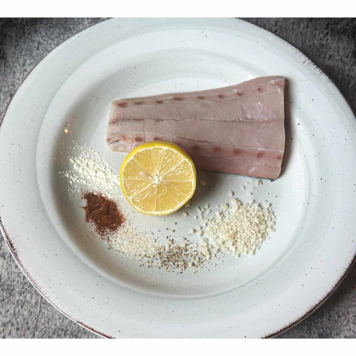 A photo representing most of the ingredients for air fryer Mahi Mahi on a plate with a Mahi Mahi fillet portion, half of a lemon, and 5 small piles of different spices, including the panko bread crumbs.