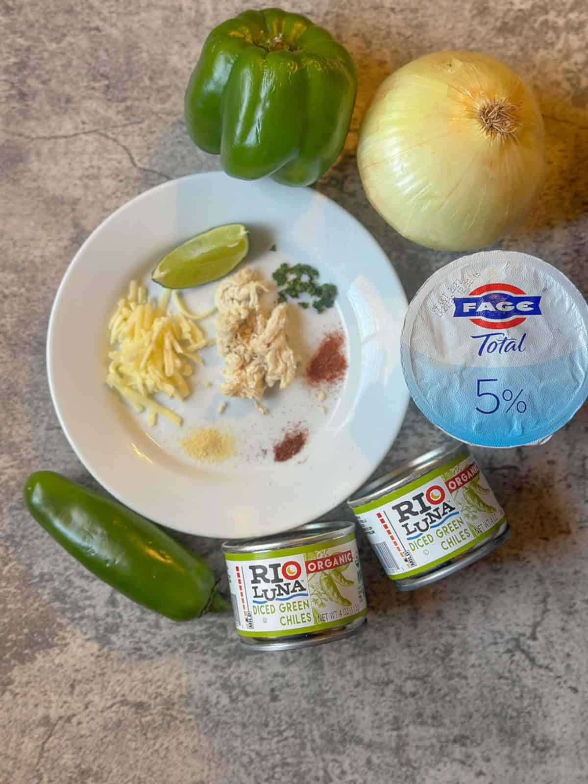 A photo of the ingredients for green chile chicken casserole. Shredded cheese, shredded chicken, a lime wedge, cumin, garlic powder, paprika, chipotle powder, and cilantro are on a white plate. around the plate is a green jalapeno, a green bell pepper, two cans of rio luna organic diced green chiles, an onion, and a small container of 5% fage yogurt.
