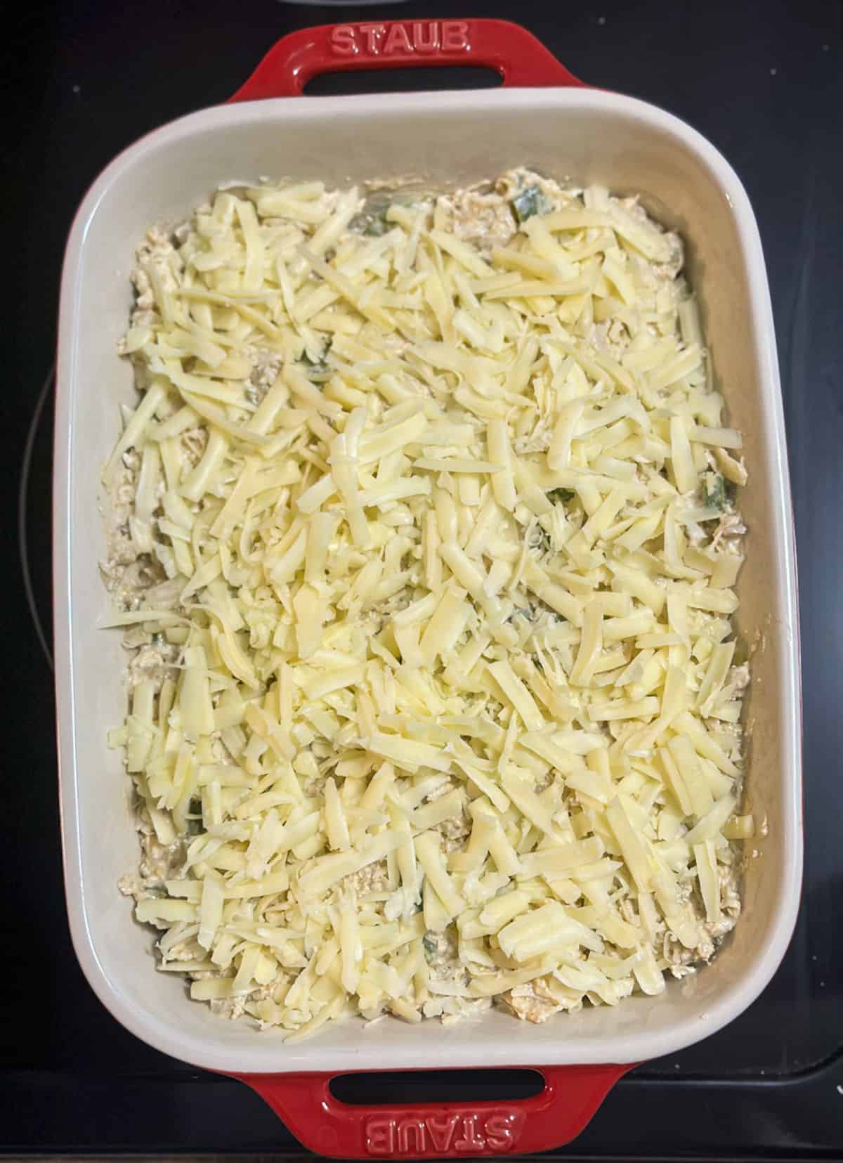 A red and white Staub casserole dish with creamy chicken and green chili mixture topped with shredded Monterey jack cheese, ready to put in the oven.