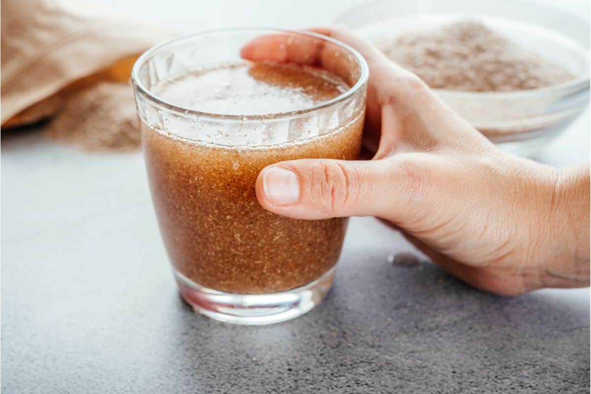 A clear glass with a man's hand holding it. in the glass is a hydrated psyllium husk drink and in the background is a bowl and a bag of psyllium husk powder.