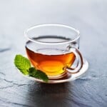 A photo of an amber colored tea in a clear glass on a white saucer with a mint leaf next to it after having sea moss gel added to it.