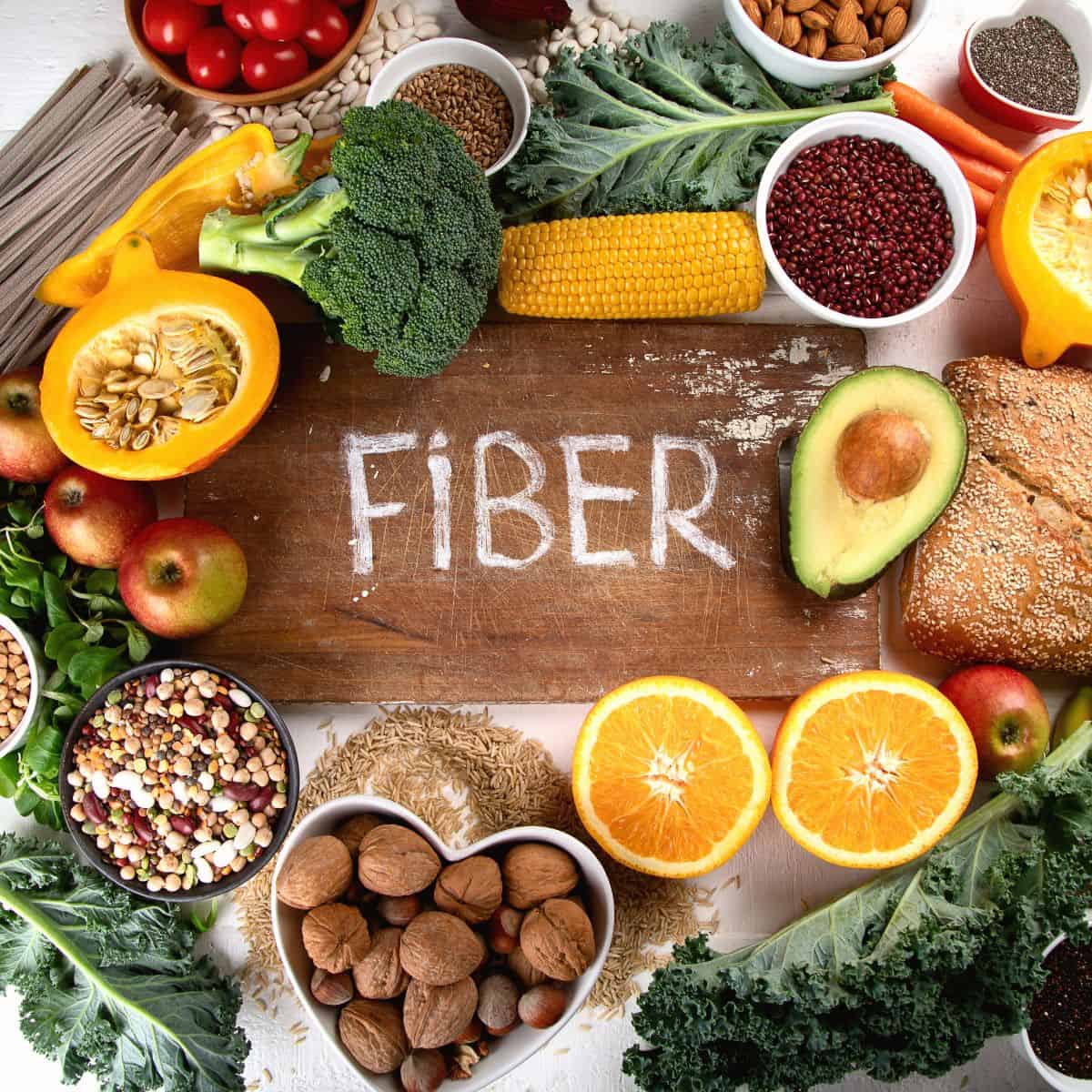 The word fiber is written in chalk on a board surrounded by various forms of fiber including leafy greens, nuts, seeds, fruit, grains, psyllium husk, and vegetables.
