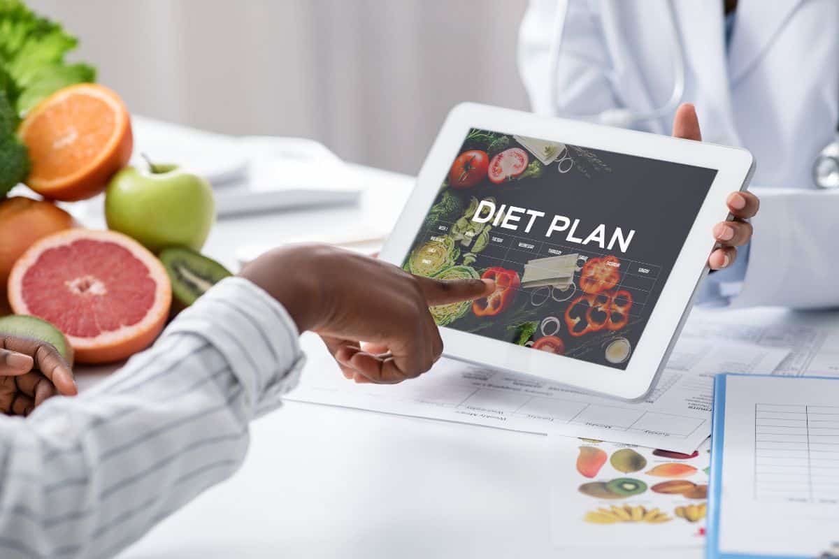 A hand is pointing toward a tablet that is held by a doctor. The tablet says diet plan with a calendar on it. around them are various fruits and vegetables and charts with food on them representing a coaching moment.