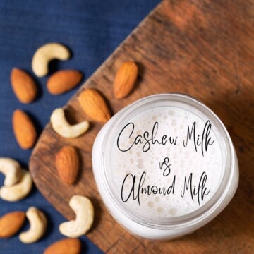 An aerial view of an open mason jar filled with milk on a dark wooden board set on a dark blue cloth surrounded by various cashews and almonds. In Cursive text inside the milk glass says Cashew Milk vs Almond Milk