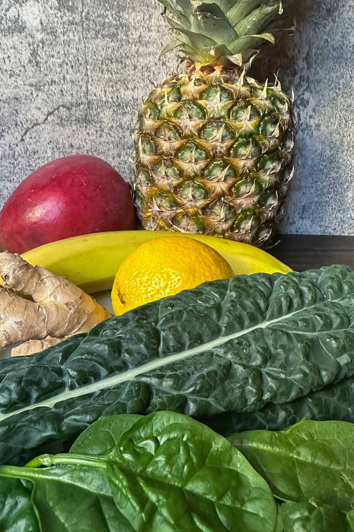 Fresh spinach, lacinto kale, ginger root, a lemon, a red mango, and a whole pineapple are shown to indicate the ingredients for the detox island green smoothie.