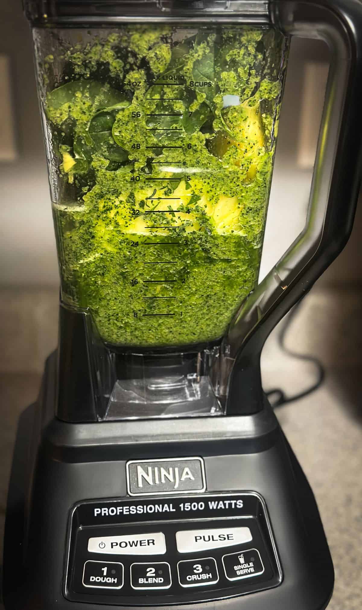 A black ninja blender filled with partially blended ingredients like spinach, kale, pineapple, and mango.