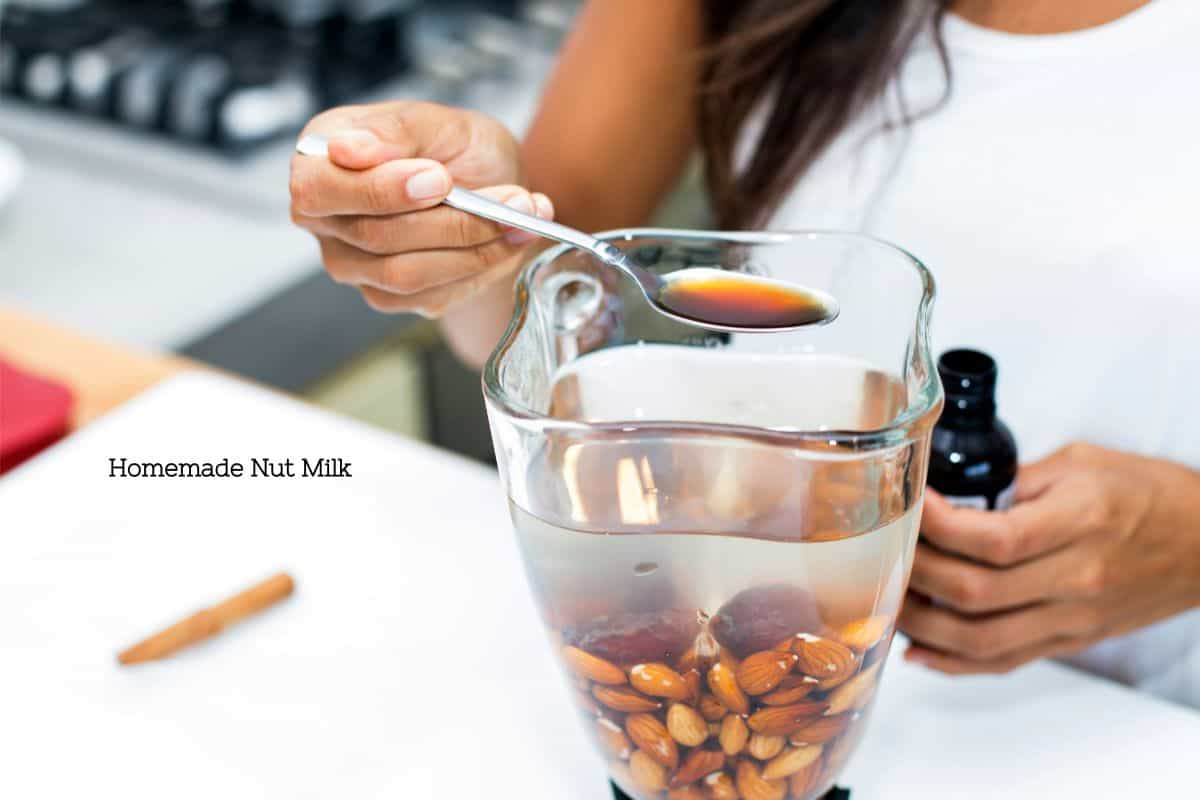 A woman standing over a blender that has almonds and water in it with a spoonful of vanilla extract about to add it in. The text overlay says "homemade nut milk"