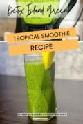 A photo of a tall glass of detox island green smoothie with bright green colors and tiny dark green speckles. The glass is garnished with a slim slice of pineapple. a transparent white box at the top says Detox Island Green. In the middle two strips of orange diagonally placed have words that say tropical smoothie recipe and at the bottom mindfullyhealthyliving.com