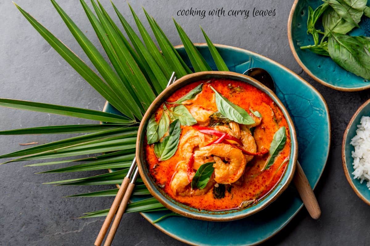 A bowl of red shrimp curry with fresh curry leaves on top, chop sticks next to it, a spoon, and decorative palm fronds.