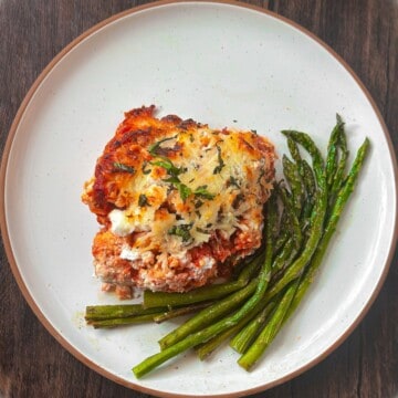 A white plate with a brown rim has a serving of keto Italian casserole with basil garnished on top, sitting next to a swirl of asparagus spears with a brown wooden background.