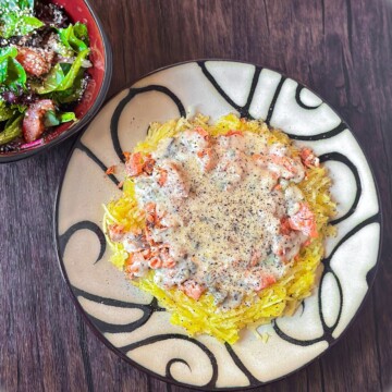 A plate of spaghetti squash topped with salmon chunks and creamy cajun alfredo sauce, seasoned with black pepper. A bowl of salad is visible in the top corner.