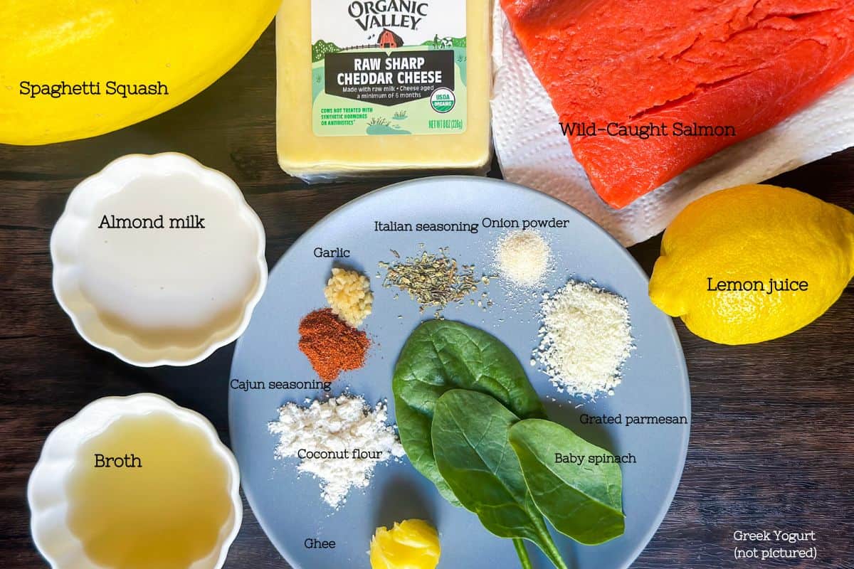 A photo of the ingredients for cajun salmon alfredo with their respective names written next to each ingredient. a spaghetti squash, a wild-caught salmon fillet, a block of raw sharp cheddar cheese, a lemon, almond milk and broth each in a ramekin with a plate in the middle with ghee, coconut four, baby spinach, cajun seasoning, garlic, Italian seasoning, onion powder, and grated parmesan to the right is lettering that says greek yogurt (not pictured)