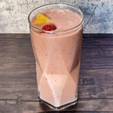 A clear glass of pink pineapple surf smoothie on display with pieces of pineapple and and strawberry on top as a garnish