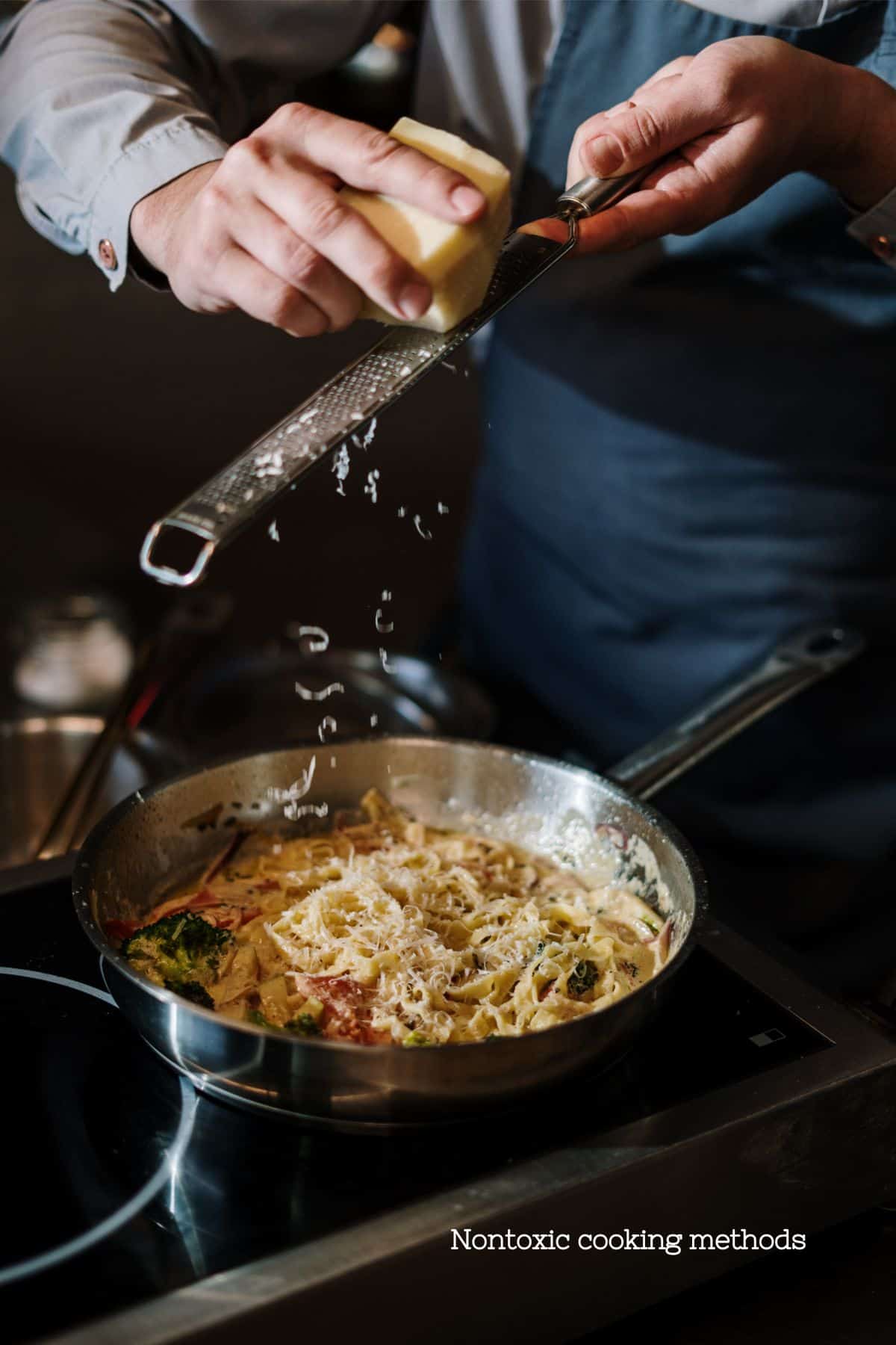 A chef grating cheese over a stainless steel pan with pasta, symbolizing non-toxic cooking methods and kitchen safety