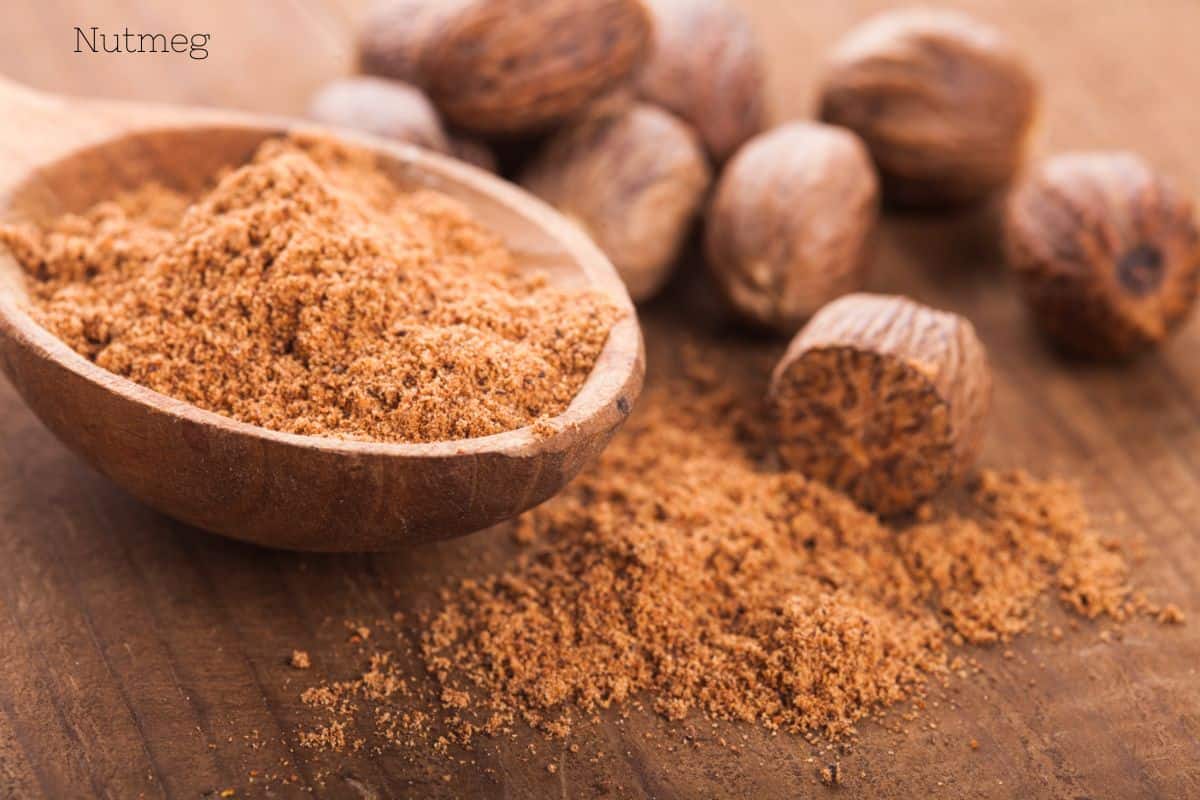 Ground nutmeg in a wooden spoon with whole nutmeg seeds scattered on a table