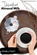 A pinterest optimized image of a brown serving tray with a white coffee mug and saucer that a woman's hands are holding. Next to it is a silver kettle and a sprig of tiny flowers. text overlay says does unsweetened almond milk in coffee break a fast? mindfullyhealthyliving.com