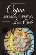 A decorative plate of spaghetti squash topped with salmon chunks and creamy cajun alfredo sauce, seasoned with black pepper. A bowl of salad is visible in the top left corner. with a brown background and white text that is elegant. It says, "cajun salmon alfredo low carb." in a box at the top says mindfullyhealthyliving.com