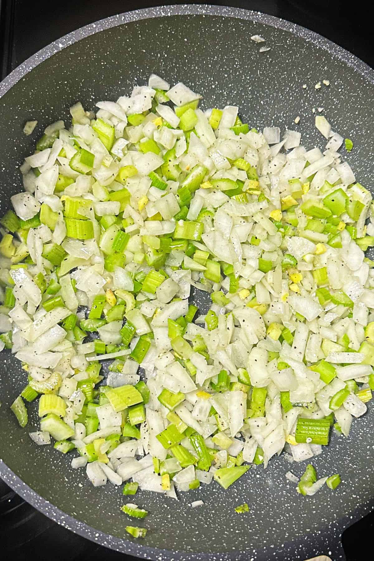 Diced celery and onions being sautéed in a skillet, seasoned with black pepper.