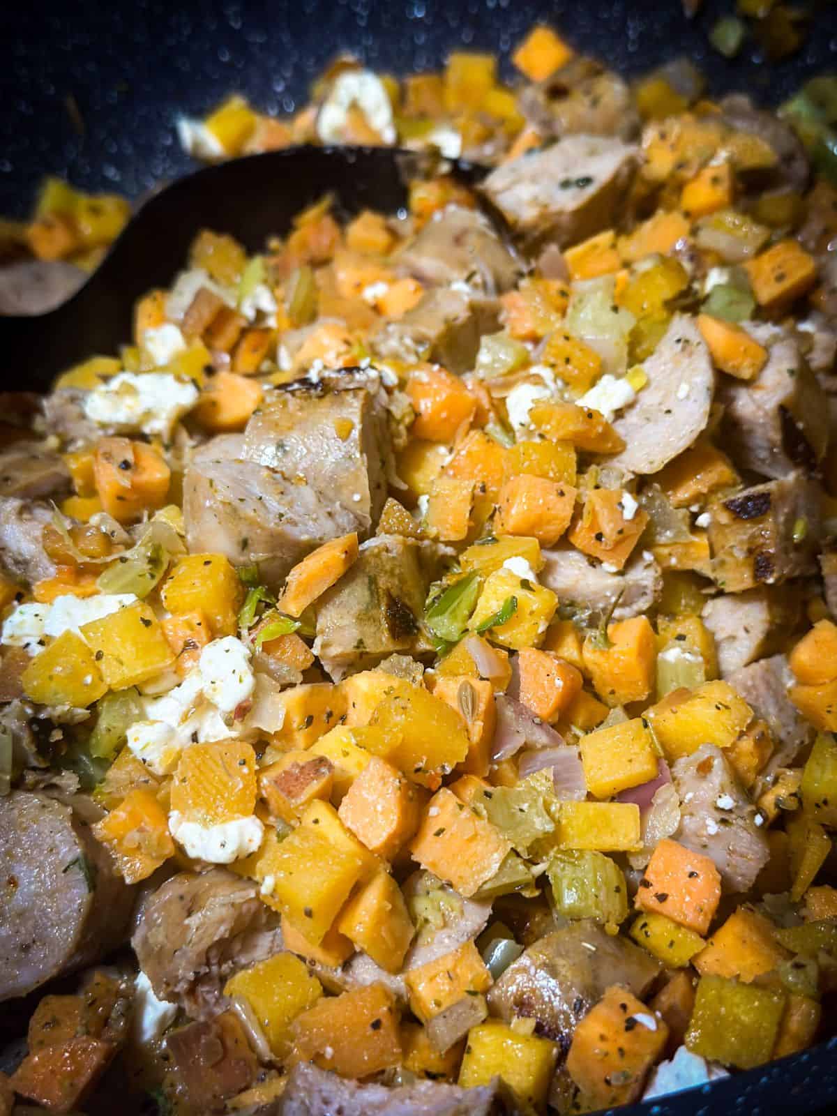 Close-up of a pan with diced sweet potatoes, celery, and onions, mixed with crumbled sausage and feta cheese, all cooked to a golden brown