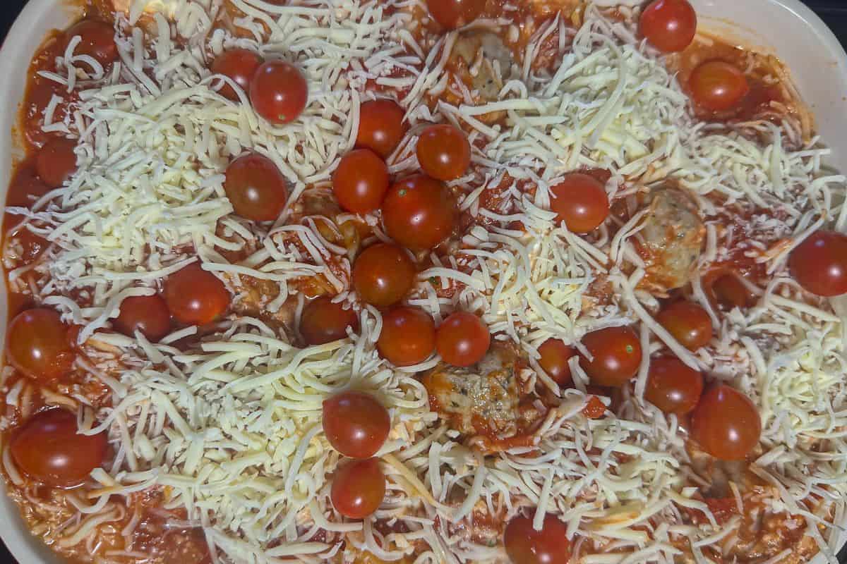 Top view of a casserole dish layered with marinara sauce, meatballs, and cherry tomatoes, ready for the next layer of cheese