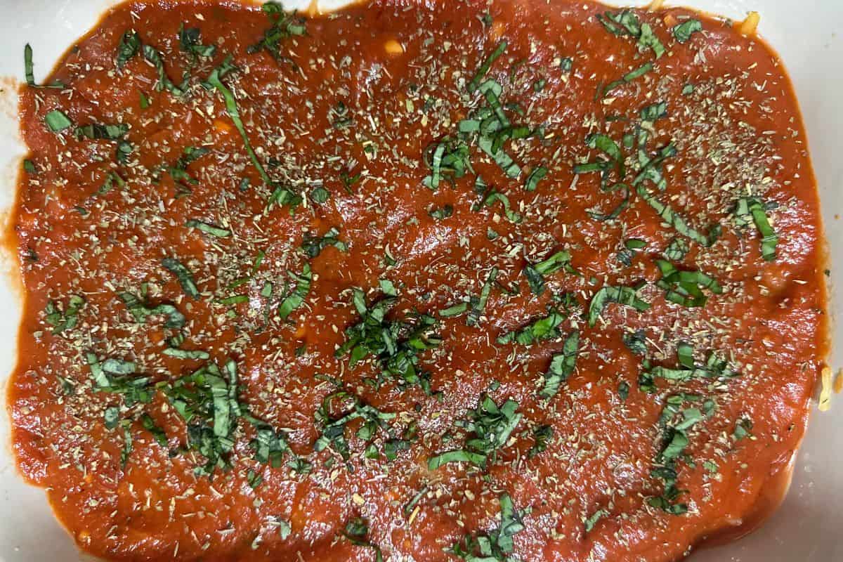 A casserole dish showing the first layer of marinara sauce sprinkled with chopped basil and Italian seasoning over the spaghetti squash