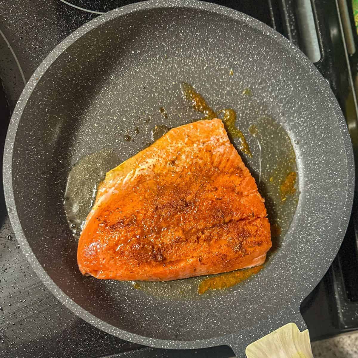 a pinkish orange salmon fillet seasoned with cajun spices in a pan with oil in it.