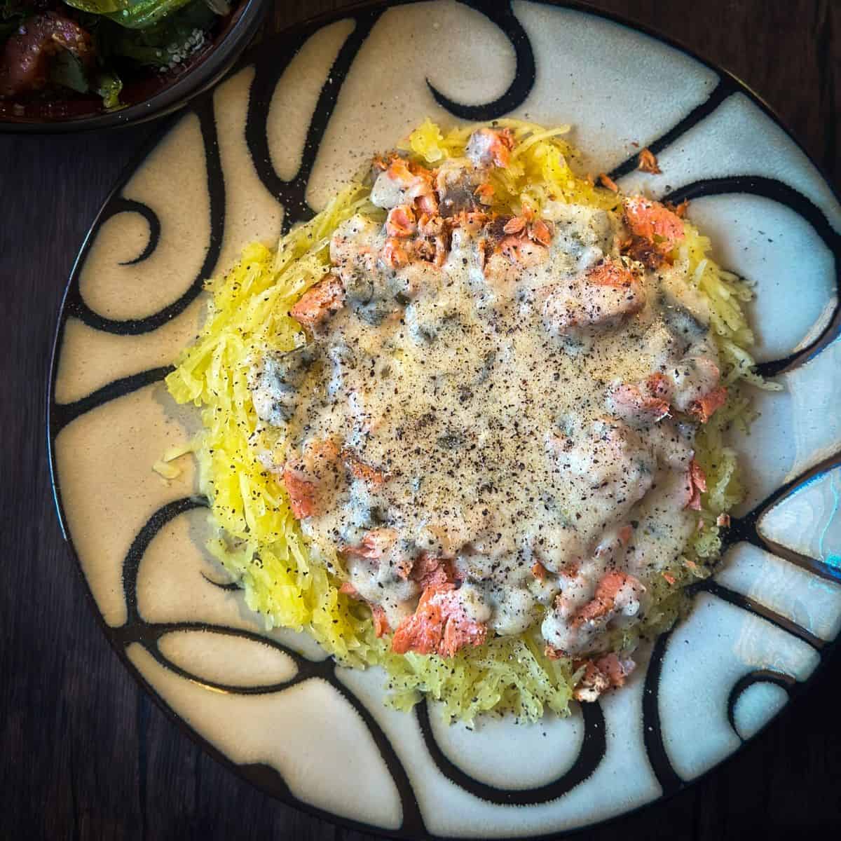 A darkened photo of a decorative plate of spaghetti squash topped with salmon chunks and creamy cajun alfredo sauce, seasoned with black pepper. A bowl of salad is visible in the top left corner.