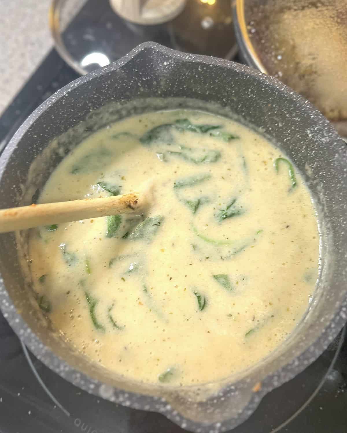 Creamy alfredo sauce cooking on a stovetop with some spices visible and wilted spinach being stirred with a wooden spoon.