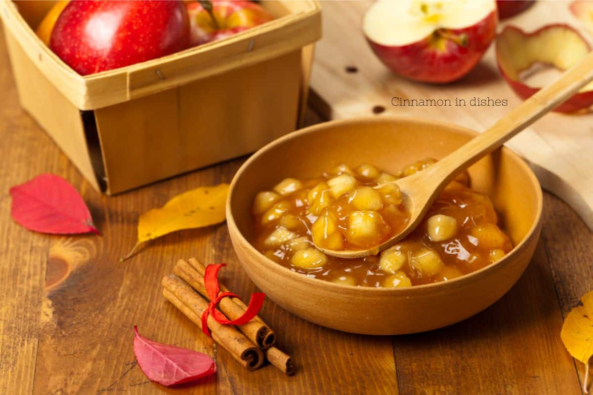 A bowl of apple cinnamon compote, with fresh apples and cinnamon sticks on a wooden table.