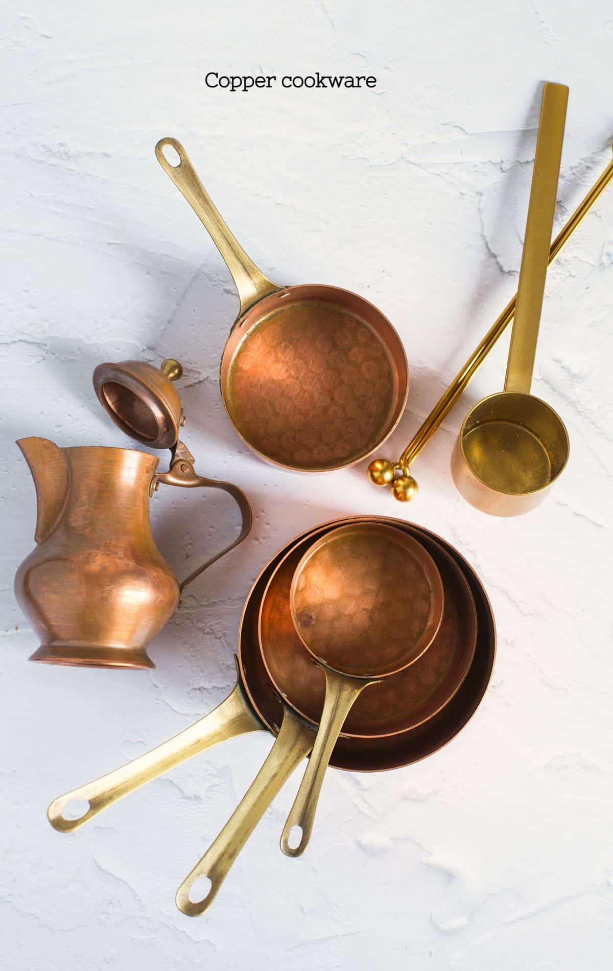 An elegant set of copper cookware, including saucepans and a ladle, with a polished finish and brass handles, arranged on a white textured background, symbolizing the risk of excessive copper leeching into foods.