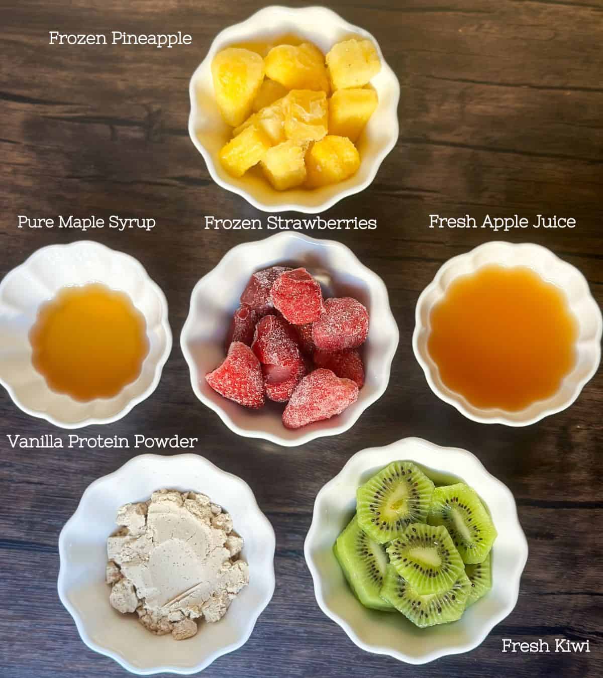 5 white ramekins in a star pattern with the ingredients in each of them for a pineapple surf smoothie with the names of the ingredients to the side written in white. The ingredients are Fresh kiwi, vanilla protein powder, frozen pineapple, frozen strawberry, pure maple syrup, and fresh apple juice.