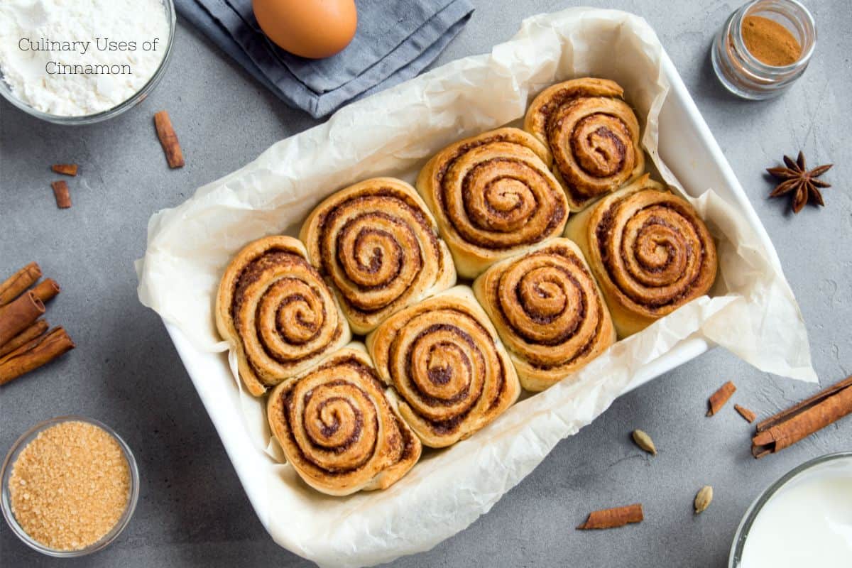 Freshly baked cinnamon rolls in a baking dish, with ingredients like cinnamon sticks nearby.