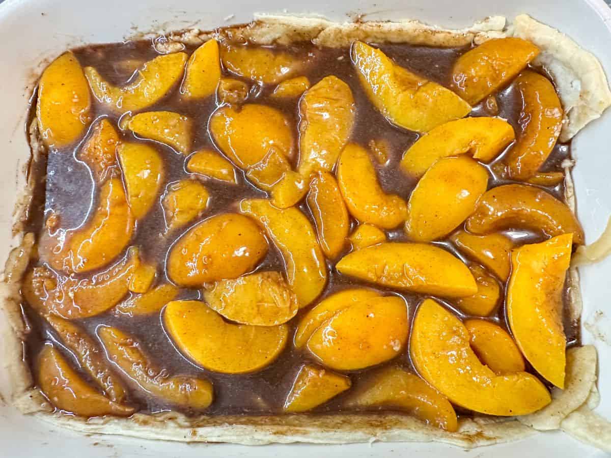 A baking dish with pie crust filled with sliced peaches and thickened brown sauce for peach cobbler.