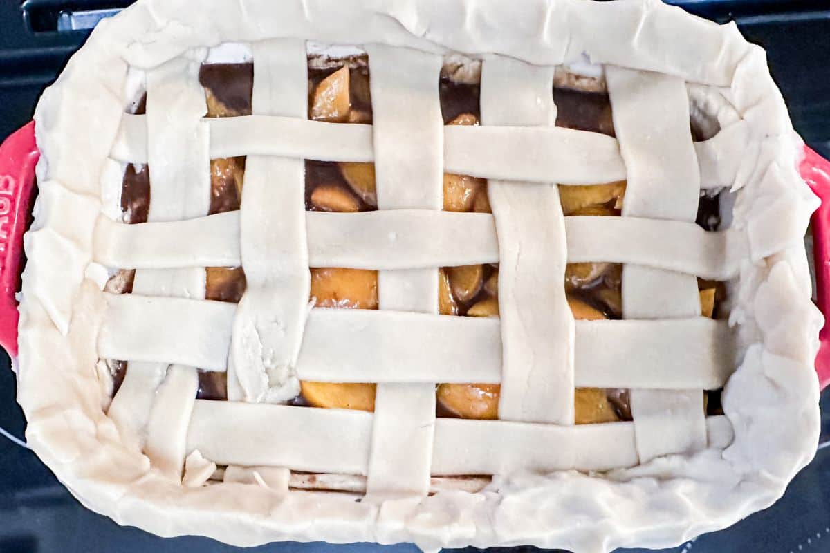 Raw pie crust strips laid atop a peach cobbler in a woven pattern, pre-baking