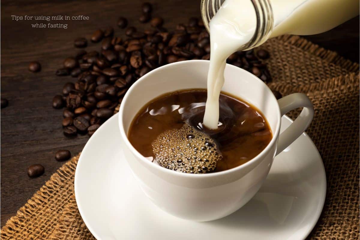 A white cup of coffee on a saucer next to a pile of coffee beans. a jug of milk is being poured into the cup. text overlay says tips for using milk in coffee while fasting.